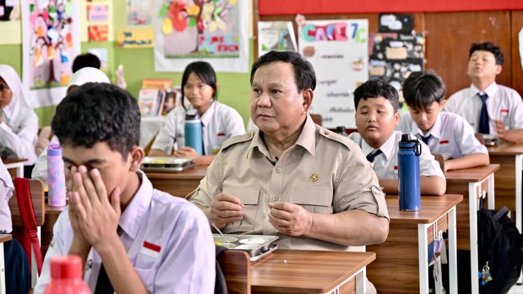 Man in a khaki uniform seated at a desk in a classroom surrounded by students in white shirts and ties